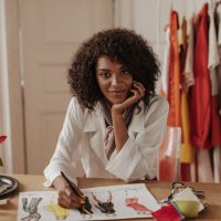 Beautiful young dark-skinned curly brunette woman in white blouse looks into camera, leans on table and designs stylish clothes.